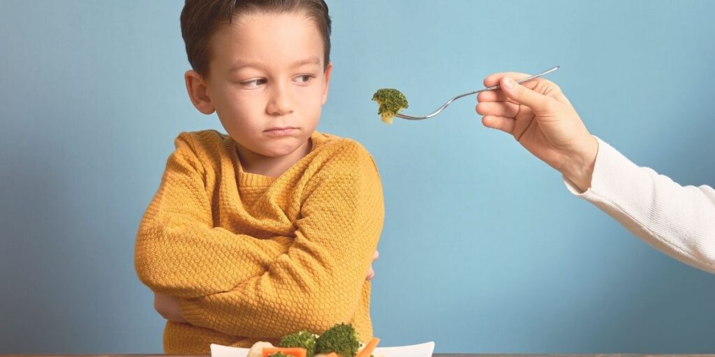 Un jeune garçon portant un pull jaune moutarde regarde avec mécontentement une fourchette tenue par une main adulte qui lui présente du brocoli. L'enfant a les bras croisés dans une posture de refus. L'image est prise devant un fond bleu clair et illustre une scène typique de difficulté alimentaire avec un enfant réticent à manger ses légumes. - Famille GAPS et santé intestinale